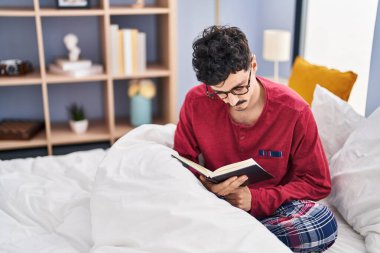 Young caucasian man reading book sitting on bed at bedroom