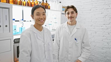 Beyaz laboratuvar önlüklü bir adam ve kadın, profesyonel bir bilimsel çevreyi resmeden, iyi donanımlı bir laboratuvarda gülümsüyor..