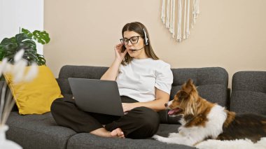 Gözlüklü ve kulaklıklı genç İspanyol kadın evcil köpeğinin yanındaki koltukta dizüstü bilgisayar kullanıyor..