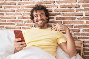 Yatağın üzerinde akıllı telefon kullanan İspanyol genç adam mutlu ve gururlu bir kendini işaret ediyor. 