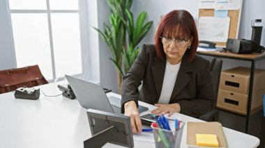 Ofis masasında bilgisayar, telefon ve belgelerle çalışan olgun İspanyol kadın..