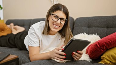 Gözlüklü, gülümseyen genç bir İspanyol kadın evdeki bir kanepede elinde tabletle rahatça yatıyor..