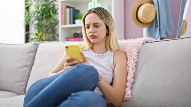 Evdeki kanepede oturan akıllı telefon kullanan genç sarışın kadın.