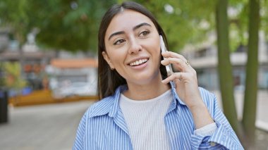 Gülümseyen genç bir İspanyol kadın şehir parkının yeşilliğinin tadını çıkarırken telefonuyla sohbet ediyor..