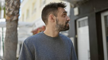 Rahat bir yaşam tarzı olan havalı ve yakışıklı İspanyol bir adam, açık havada ciddi bir ifadeyle çekilmiş, güneşli bir şehir sokağına bakıyor..