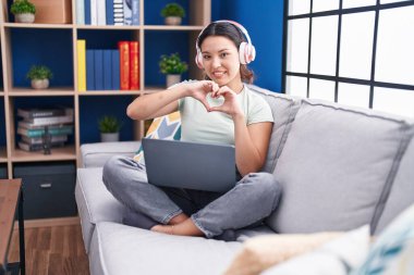 Evde dizüstü bilgisayar kullanan, kulaklık takan, kalp sembolü ve elleri ile gülümseyen İspanyol genç bir kadın. romantik kavram. 