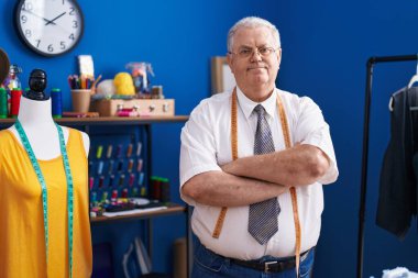 Orta yaşlı, gri saçlı, sırıtan, kendine güvenen terzi terzide kollarını kavuşturup el kol hareketi yapan bir adam.