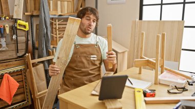 Genç sakallı bir adam, ahşap kalaslar ve aletlerle çevrili bir marangoz stüdyosunda çalışıyor..