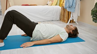 Genç sakallı bir adam, modern dekorlar ve ev bitkileriyle dolu parlak bir yatak odasındaki yoga minderinde dinleniyor. Huzur ve sükunetin vücut bulmuş hali..