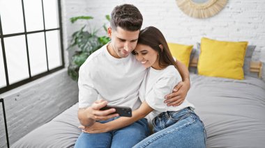 Sevgi dolu bir çift, evlerinde samimiyet ve teknoloji tasvir eden modern yatak odalarında akıllı bir telefona bakarken kucaklaşır..