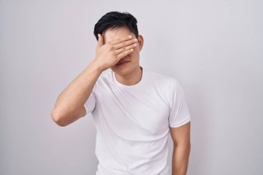 Genç Asyalı bir adam beyaz arka planda duruyor. Elle gözlerini kapatıyor, ciddi ve üzgün görünüyor. Görme, saklama ve reddetme konsepti 