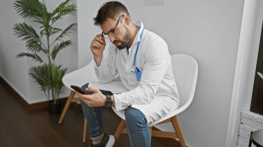 Yakışıklı bir İspanyol genç adam, odaklanmış bir doktor, klinik bekleme odasında otururken akıllı telefonuyla mesaj atıyor, işine dalmış, modern tıp profesyonelliğinin bir portresi..