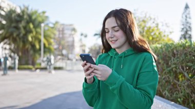 Güneşli bir şehir parkında akıllı telefon kullanan gülümseyen genç bir kadın.