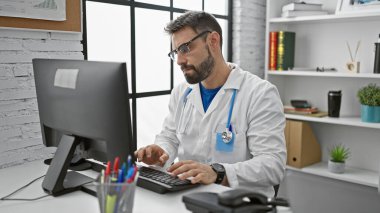 Sakallı, genç, İspanyol bir doktor, bir klinikte bilgisayarını kullanıyor.