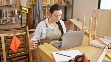 İspanyol kadın, dizüstü bilgisayara bakarken marangozluk atölyesinde ahşabı ölçüyor.