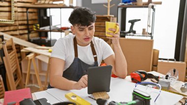 Genç bir adam, iyi donanımlı bir marangoz atölyesinde elinde bardak tutarken tabletini inceliyor..