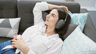 Rahat genç bir kadın, rahat bir oturma odasında, elinde akıllı bir telefonla koltukta uzanırken kulaklıkla müzik dinlemekten hoşlanır..