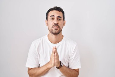 Beyaz arka planda duran yakışıklı İspanyol adam. Elleriyle yalvarıyor ve dua ediyor. Yüzünde umut ifadesi var. Yalvarıyor. 