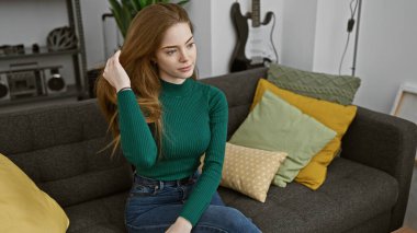 Sarışın, beyaz bir kadın, renkli yastıklarla bezenmiş çağdaş bir kanepede rahatlar..