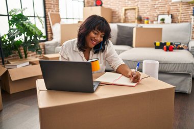 Young beautiful latin woman talking on smartphone writing on notebook at new home
