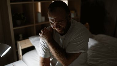 Omzu ağrıyan, üzgün ve endişeli beyaz bir adam, sabahları yatakta, hayal kırıklığı içinde yaralı omzuna dokunuyor..