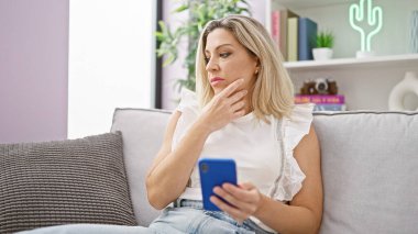 Genç sarışın kadın akıllı telefon kullanıyor. Kanepede oturuyor ve evde oturup düşünüyor.