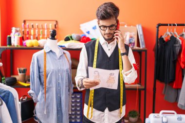Young hispanic man tailor talking on smartphone looking clothing design at atelier