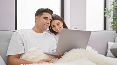 Bir kadın ve bir erkek evlerinde kucaklaşıp rahat bir kanepede dizüstü bilgisayarlarını paylaşıyorlar. Sevgi ve birliktelik hissi yaratıyorlar..