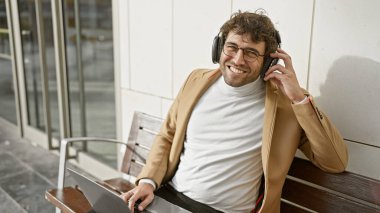 Gözlüklü ve kulaklıklı neşeli genç bir adam şehirde bir bankta otururken dizüstü bilgisayarında müzik dinlemekten hoşlanıyor..