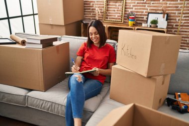 Young beautiful hispanic woman smiling confident writing on notebook at new home