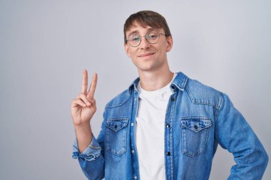 Beyaz, sarışın, gözlüklü, gülümseyen, parmaklarını zafer işareti yaparken gösteren bir adam. İki numara.. 