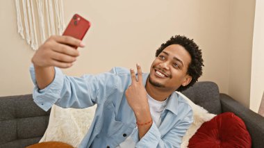 Gülümseyen adam kanepenin içinde kırmızı telefonuyla selfie çekiyor. Barış işareti ve günlük tarzını gösteriyor..