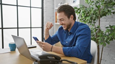 Ofiste kutlama yapmak için dizüstü bilgisayar ve akıllı telefon kullanan İspanyol bir iş adamı.
