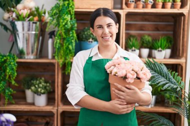 Young beautiful hispanic woman florist smiling confident holding plant at flower shop