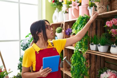 Young beautiful arab woman florist using touchpad holding plant of shelving at flower shop