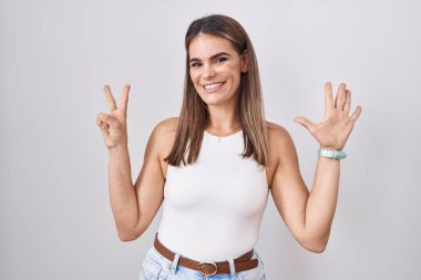 Beyaz arka planda duran Latin kökenli genç bir kadın yedinci parmaklarını gösterip işaret ederken kendinden emin ve mutlu gülümsüyor.. 