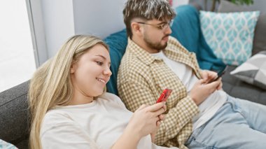 Genç çift rahat bir oturma odasındaki akıllı telefonları kullanarak kanepede uzanıyorlar..