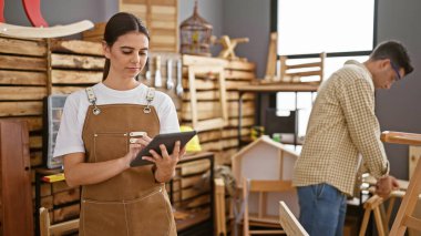 İyi donanımlı bir marangoz stüdyosunda, dijital tabletle odaklanmış bir kadın ve ahşap mobilyaları zımparalayan bir adam dinamik bir marangozluk takımı oluşturuyorlar..