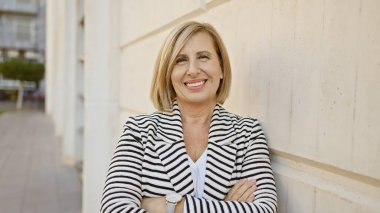 Kendine güveni tam, orta yaşlı, kollarını kavuşturmuş bir sarışın kadın. Şehir caddesinde durmuş kameraya gülümsüyor..