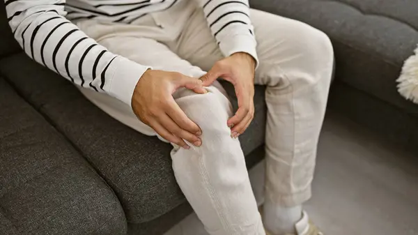 Hispanic man\'s hands gripping knee in pain, sitting on comfy living room sofa, suffering from leg joint injury at home, basking in sunlight yet unable to relax due to discomfort