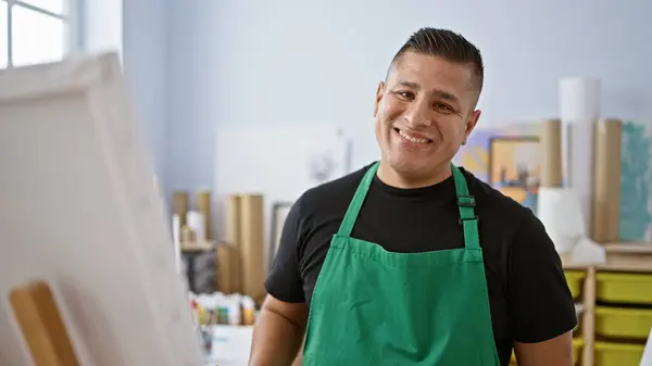 Confident young latin man artist, apron-adorned and brush in hand, standing proudly in his art studio, smiling with creative joy while drawing on a canvas