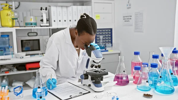 In the heart of the lab, attractive african american woman scientist seriously taking notes, working with microscope in bustling medical research center