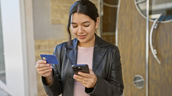 A young hispanic woman checks her credit card and smartphone on a city street, embodying urban elegance and connectivity.