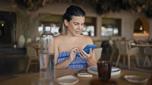 Young hispanic woman smiling happy using smartphone sitting on the table at the restaurant