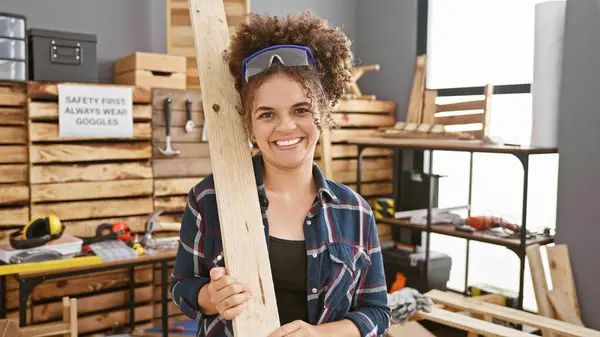 身穿安全护目镜的卷发微笑的女人在一个有组织的木工车间持木 — 图库照片
