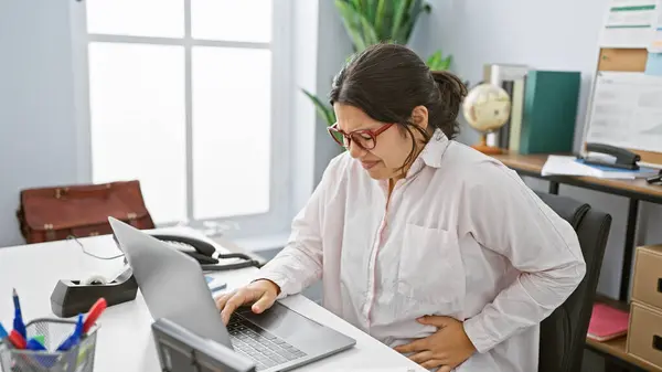 一位年轻的惊慌失措的女人带着笔记本电脑坐在办公桌前 在办公室里感到肚子痛 — 图库照片