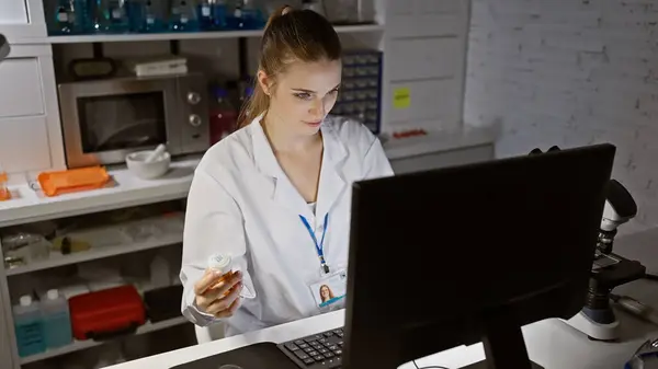 Skupiona Młoda Kobieta Pracująca Przy Komputerze Laboratorium Trzymająca Butelkę Tabletkach — Zdjęcie stockowe