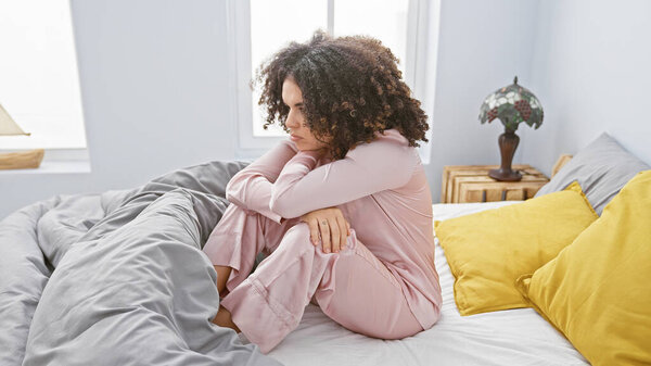 Латиноамериканская кудрявая молодая женщина в задумчивой позе сидит на кровати в ярко освещенной спальне.