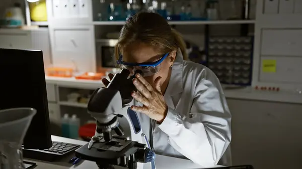 A woman examines samples under a microscope in a laboratory, wearing safety glasses and a lab coat.