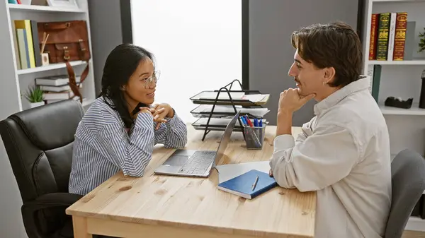 stock image A woman and a man, likely coworkers, engage in conversation at an office with a laptop and notepad on the desk.